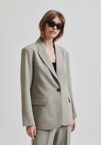 Lys grå skreddersydd blazer med en enkelt svart knapp, notched lapeller og en slim fit. Har brystlomme og strukturerte skuldre.