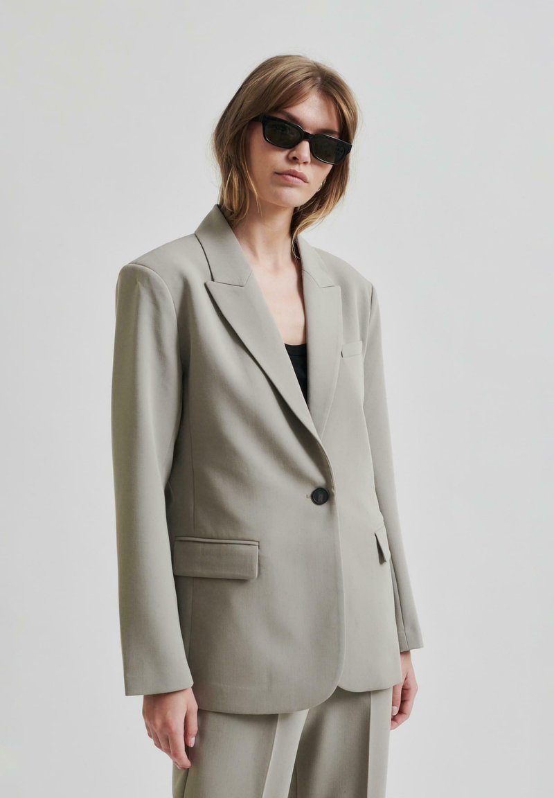 Lys grå skreddersydd blazer med en enkelt svart knapp, notched lapeller og en slim fit. Har brystlomme og strukturerte skuldre.