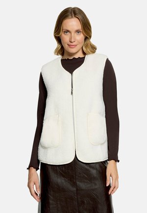 Femme aux cheveux blonds courts portant un gilet polaire blanc zippé sur un haut noir à manches longues et une jupe noire, debout devant un fond uni.