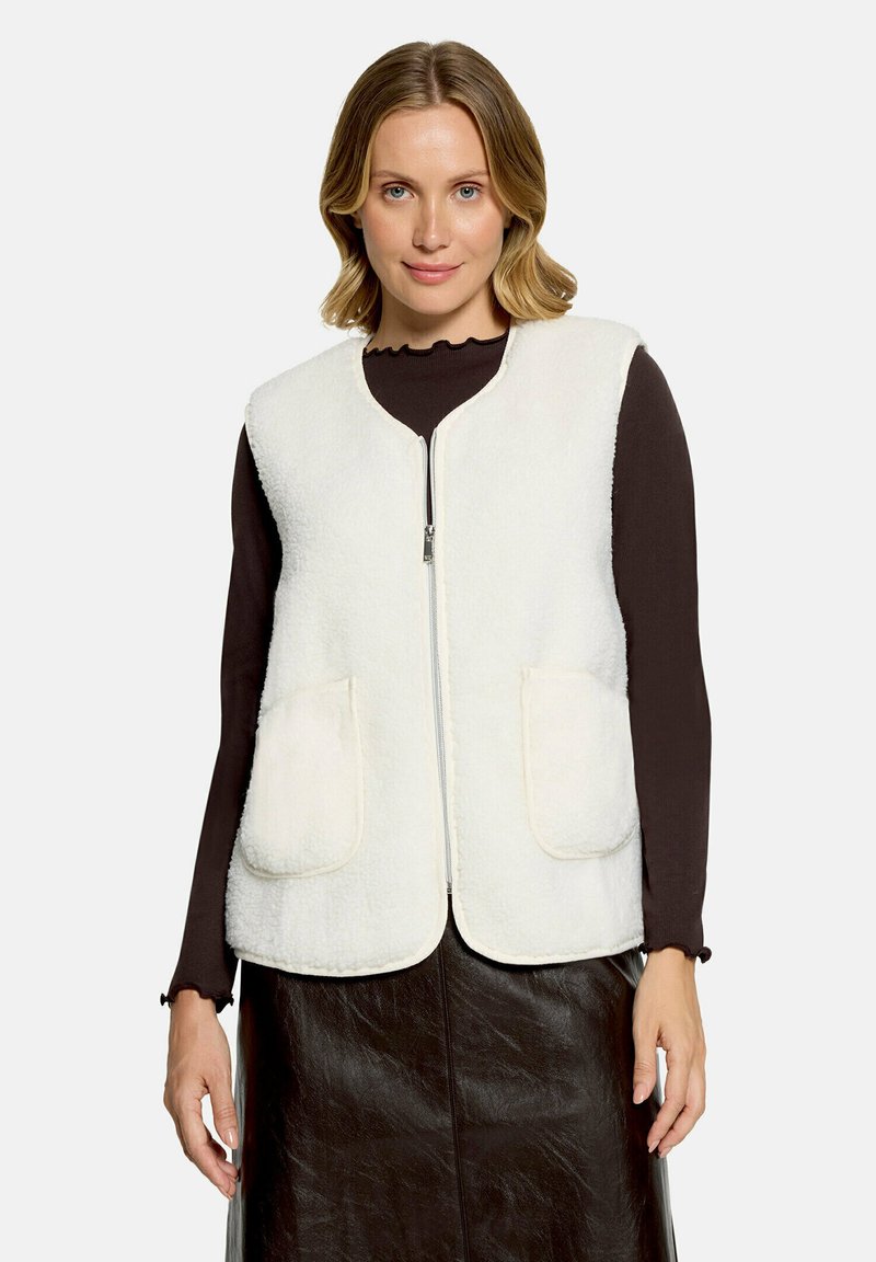 Femme aux cheveux blonds courts portant un gilet polaire blanc zippé sur un haut noir à manches longues et une jupe noire, debout devant un fond uni.