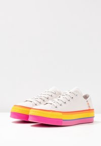 Canvas Sneaker mit einer strukturierten weißen Oberseite, die eine mehrfarbige Sohle in Pink, Gelb und Orange aufweist, mit silbernen Ösen und Schnürsenkeln.