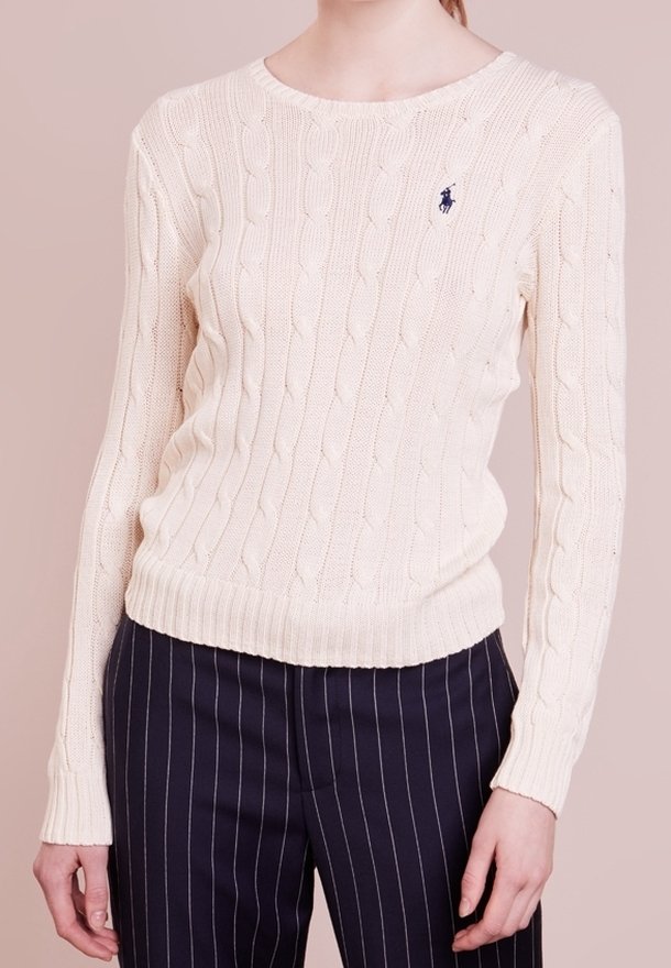 Femme portant un pull en maille torsadée crème avec un petit logo et un pantalon à rayures noires, debout devant un fond beige.