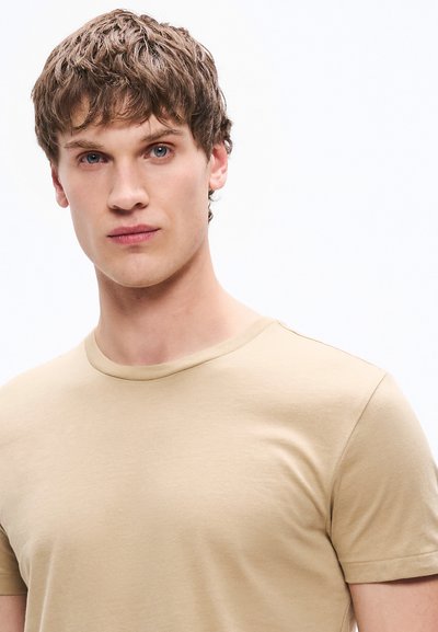 Ανοιχτό μπεζ κοντομάνικη μπλούζα με στρογγυλή λαιμόκοψη, κατασκευασμένη από απαλό ύφασμα, με λεία υφή και εφαρμοστή σιλουέτα.