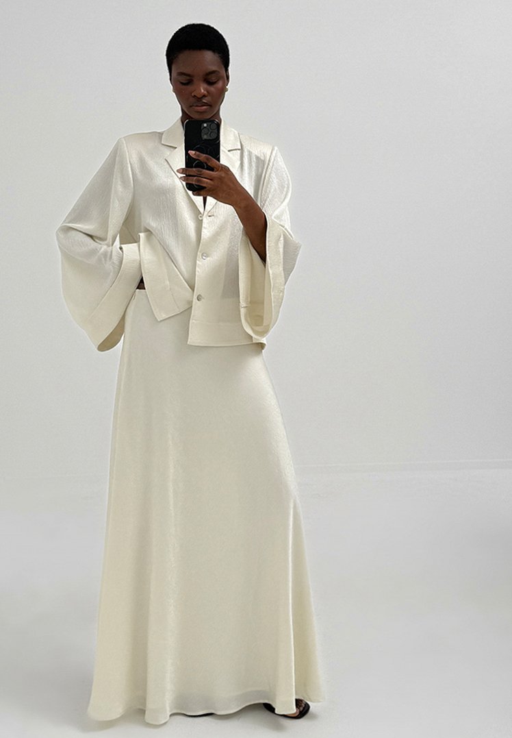 Moteris dėvi ilgą baltą satininę sijoną ir derančią švarkelį su plačiais varpelių formos rankovėmis, laiko telefoną, kad nufotografuotų veidrodinę asmenukę.