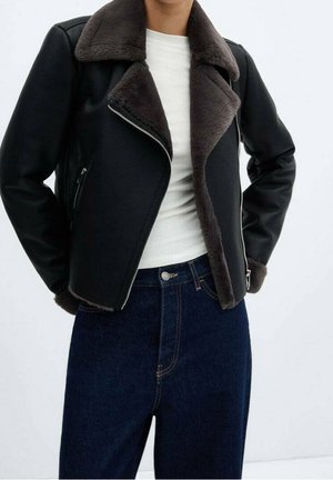 Personne portant une veste en shearling noire avec un grand col, un haut blanc et un jean taille haute bleu foncé, les mains dans les poches de la veste.