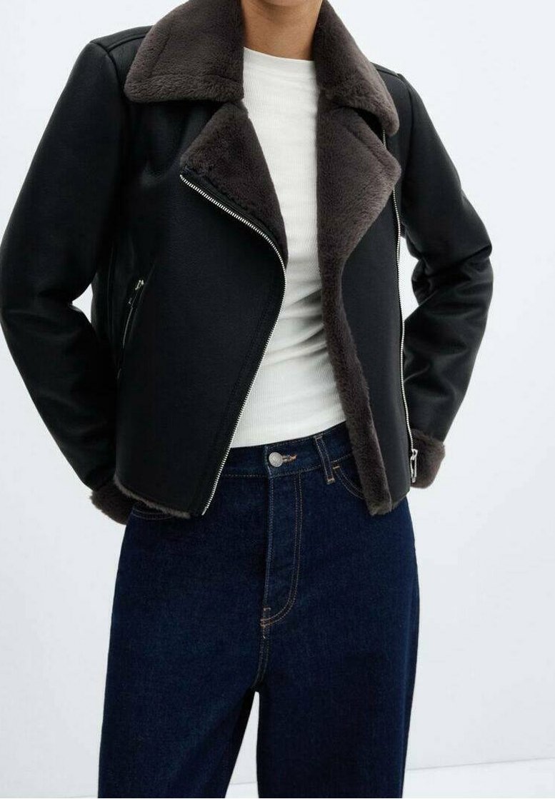 Personne portant une veste en shearling noire avec un grand col, un haut blanc et un jean taille haute bleu foncé, les mains dans les poches de la veste.