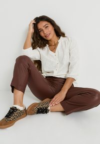 Blusa blanca texturizada con botones, pantalones marrones ajustados y zapatillas color beige con estampado de leopardo y acentos de encaje negro.