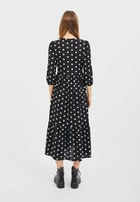 Zwarte jurk met witte polkadots, een geplooide zoom, driekwart mouwen en een getailleerde taille, gemaakt van lichtgewicht stof. Draagt zwarte enkellaarzen.