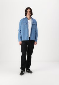 Lichtblauwe denim shirt met twee borstzakken, wit t-shirt met ronde hals, zwarte broek en zwarte sportschoenen met witte strepen.
