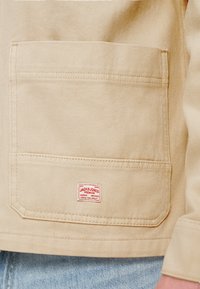 Μπεζ τζιν μπουφάν με μια μεγάλη μπροστινή τσέπη και μια κόκκινη ετικέτα με λευκό κείμενο. Διακριτά ραφές και υφή υφάσματος εμφανείς.