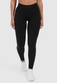 Leggings neri a vita alta con una texture liscia, design aderente e senza modelli visibili, abbinati a scarpe da ginnastica bianche.