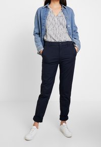 Kurtka dżinsowa w niebieskim odcieniu, wzorzysta koszula w białe i granatowe elementy oraz ciemne, tapered spodnie. Białe sneakersy dopełniają stylizację.