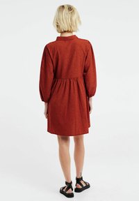 Červené šaty s černými puntíky, s límečkem, balónovými rukávy a elastickým pasem. Spodní okraj je nad kolenem. Oblečeno s černými sandály.