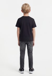 Vaalea lapsi seisoo selin, pukeutuneena mustaan t-paitaan, haalistuneisiin mustiin farkkuihin ja mustavalkoisiin tennareihin valkoisella taustalla.