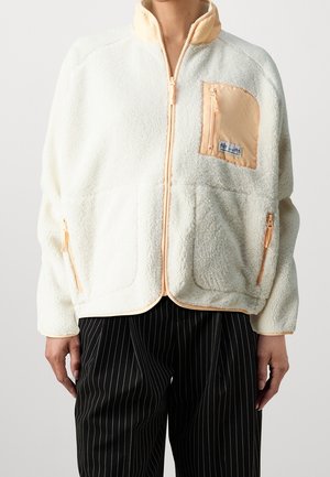 Personne portant une veste polaire blanche avec des bordures pêche et des poches zippées, associée à un pantalon noir à fines rayures.