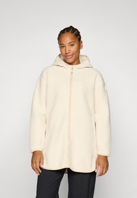 Lächelnde Frau in einer cremefarbenen Fleecejacke mit Kapuze, die vor einem hellgrauen Hintergrund steht. Lässige und entspannte Pose.