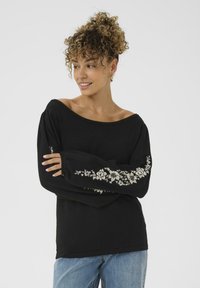 Donna con capelli ricci che indossa un maglione nero a spalle scoperte con ricami floreali bianchi sulle maniche e jeans azzurri chiari, sorridente e che guarda a sinistra.