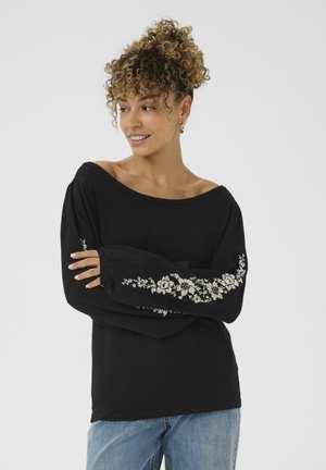 Donna con capelli ricci che indossa un maglione nero a spalle scoperte con ricami floreali bianchi sulle maniche e jeans azzurri chiari, sorridente e che guarda a sinistra.