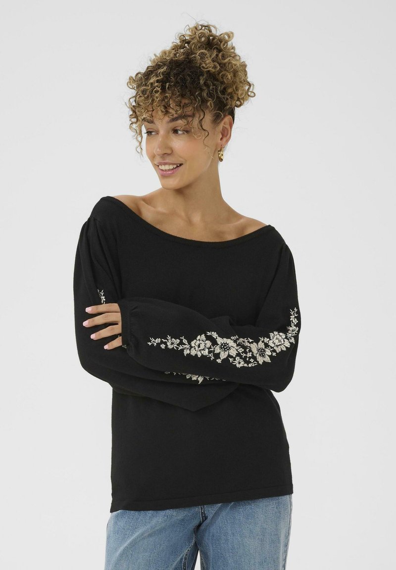 Donna con capelli ricci che indossa un maglione nero a spalle scoperte con ricami floreali bianchi sulle maniche e jeans azzurri chiari, sorridente e che guarda a sinistra.