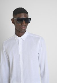 Camisa blanca de manga larga y botones de tejido ligero. Modelo que lleva unas grandes y angulares gafas de sol negras con lentes reflectantes.