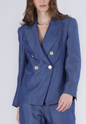 Mujer con un blazer azul entallado cruzado con botones dorados y pantalones a juego, mostrada desde el cuello hasta las caderas.