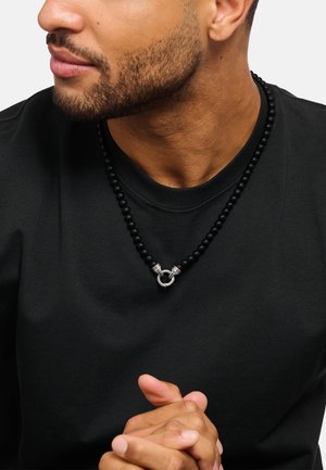 Schwarze Perlenkette mit einem runden silbernen Verschluss, getragen über einem schwarzen Rundhalsshirt. Verfügt über eine glatte Textur und ein schlichtes Design.