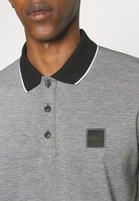 Homme portant un polo gris avec un col noir et trois boutons noirs, avec un patch carré noir portant le texte « BOSS » sur la poitrine.