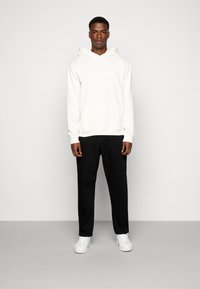 Felpa bianca con tasca frontale e logo, abbinata a pantaloni neri e sneakers bianche. Tessuto morbido, vestibilità rilassata, design semplice.