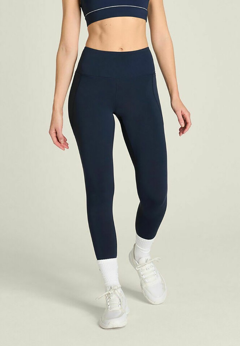 Frau trägt marineblaue Leggings mit hoher Taille, weiße Socken und weiße Sneakers, steht vor einem schlichten hellen Hintergrund.