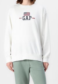 Sweatshirt blanc à col rond en coton, avec un logo "GAP" brodé accompagné d'un drapeau et de "EST. 1969" en rouge et marine. Coupe décontractée.