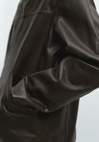 Nahaufnahme eines dunkelbraunen Lederjackenärmels mit der Hand in der Seitentasche, die die glatte Textur und die Details der Naht zeigt.