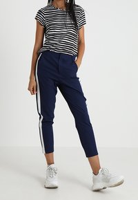 Persona vestida con pantalones tapered azul marino con franjas laterales blancas, una camiseta de rayas en blanco y negro, y zapatillas blancas, de pie con una mano en el bolsillo.