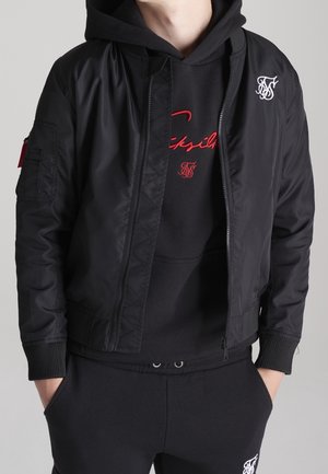 Osoba ubrana w czarną kurtkę typu bomber na czarnej bluzie z kapturem z czerwono-białymi haftowanymi logo, ręce w kieszeniach na jednolitym tle.