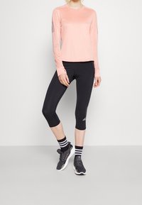 Top sportivo a maniche lunghe rosa con pannelli in rete, abbinato a leggings capri neri. Il modello indossa sneaker nere e calze alla caviglia a righe.