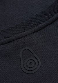 Tessuto nero con una consistenza liscia, caratterizzato da un logo circolare con finitura opaca. Collo a costine con un design aderente.