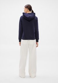 Námořnicky modrá mikina s měkkou texturou, s velkou kapucí a těsnými manžetami, doplněná světle barevnými širokými kalhotami s kapsami na zadní straně.