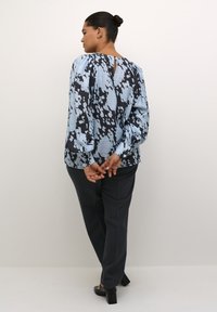 Hellblaue Bluse mit einem schwarzen abstrakten Muster, langen Ärmeln und einem Schlüssellochdetail im Rücken. Kombiniert mit dunklen Hosen und Blockabsätzen.