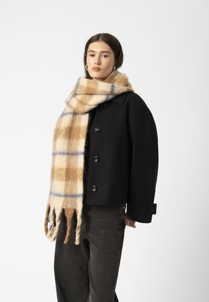 Kvinde iført en sort knapjakke, lang beige og blå ternet tørklæde, mørke bukser, stående mod en ensfarvet lys baggrund.