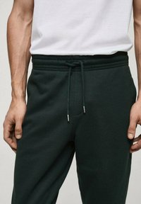 Temnozeleno trenirko hlače z vrvico v pasu, ki vsebujejo srebrne kovinske konice. Material izgleda mehko, z gladko teksturo in elastičnim pasom.