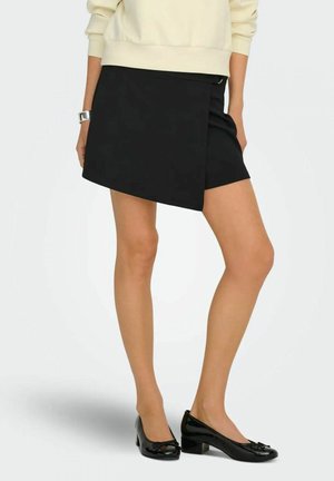 Donna che indossa una felpa color crema, gonna avvolgente asimmetrica nera, braccialetto d'argento e scarpe nere in vernice con tacco basso.