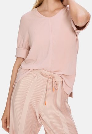 Kvinde iført løs, pastel lyserød kortærmet skjorte stoppet ned i brede, teksturerede bukser med snor i taljen.