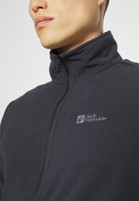 Schwarze Fleecejacke mit hohem Kragen, Reißverschluss und gesticktem Logo auf der Brust. Weiche Textur und taillierte Passform.