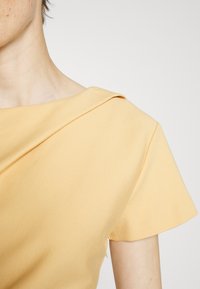 Mosterdgele top met een brede halslijn en korte mouwen, met een gladde stof en subtiele drapering op de schouder.