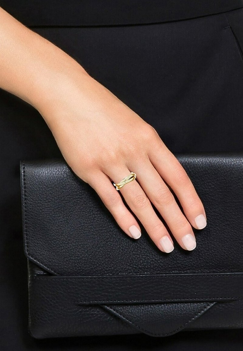 Main avec des ongles nus polis portant une bague torsadée en or, reposant sur une pochette en cuir noir texturé sur un fond noir.
