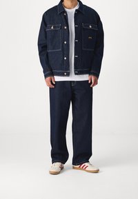 Tmavě modrá džínová bunda s dvěma náprsními kapsami, zapínáním na knoflíky a bílým šitím, doplněná volnými tmavými džínami a béžovými botami s červenými pruhy.