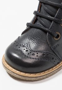 Scarpa di pelle nera con punta arrotondata, dettagli brogue e cuciture marroni. Presenta lacci e una suola in gomma testurizzata.