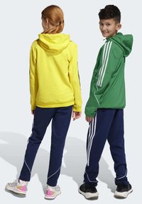 Geel hoodie met een trekkoord, gecombineerd met marineblauwe joggers. Groene jas met drie witte strepen op de mouwen, gedragen met zwarte sneakers.