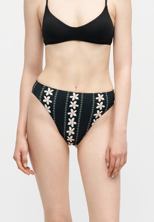 Fekete bikiniben lévő nő, fehér és rózsaszín virágos csíkos mintával a bikini alsó részén, és egyszínű fekete bikini felsővel.