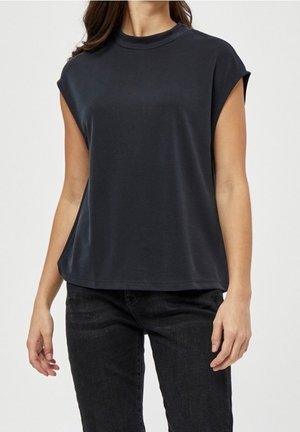 Kvinde iført løs, ærmeløs sort top og sorte jeans, stående med armene afslappet ned langs siderne mod en ensfarvet baggrund.