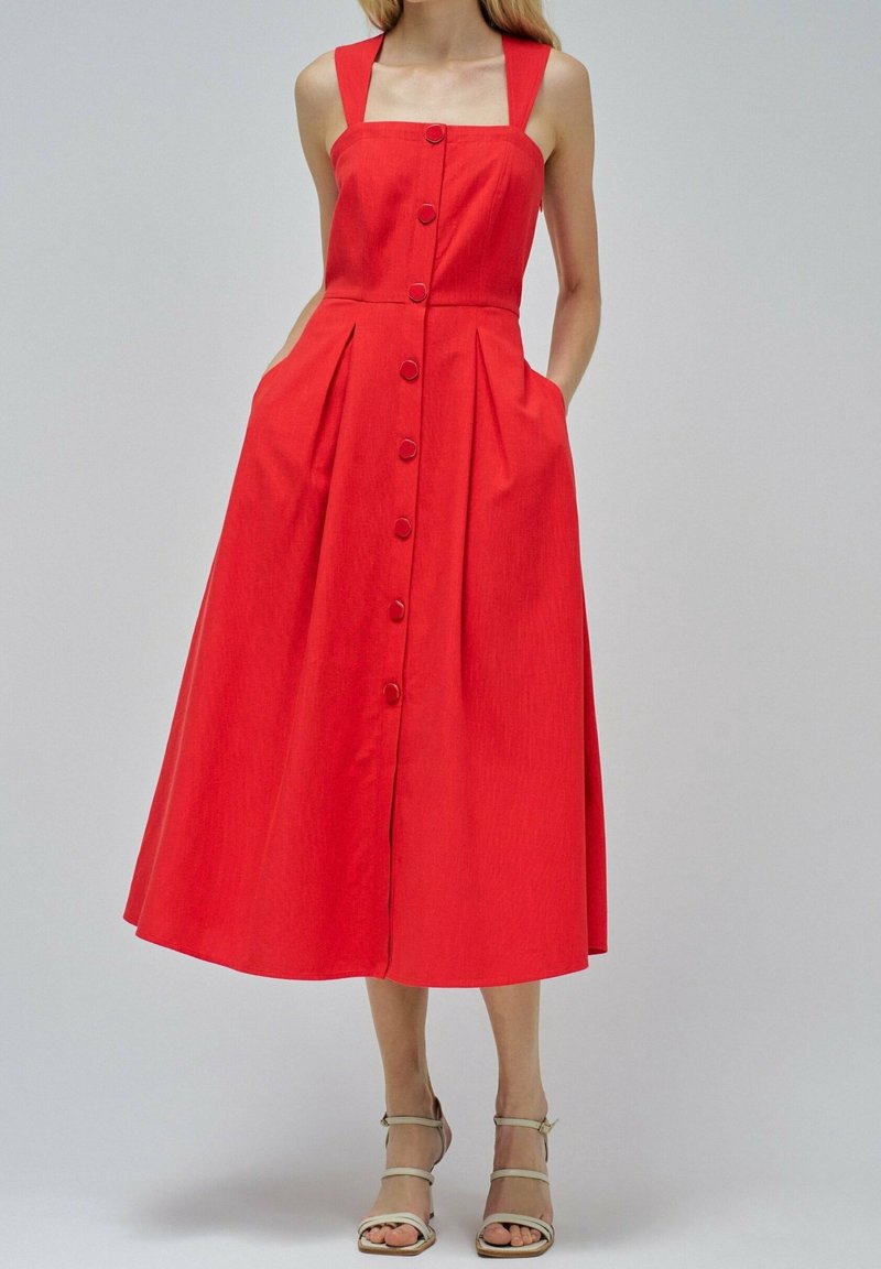 Femme portant une robe midi rouge sans manches à boutons sur le devant, avec larges bretelles et poches, associée à des sandales à talons ouvertes beiges.